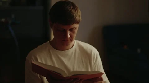 Deeply concentrating young man reading paper book while sitting on cozy sofa in Stock Footage 314269001
