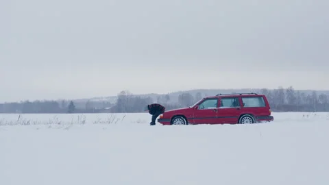 Dejected man sitting on hood of broken down red car on snowy country road Stock Footage 331756443