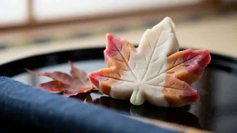 A delicate maple leaf shaped wagashi sweet sits on a dark plate next to a real Stock Photos