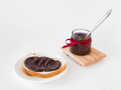 Delicious chocolate spread on bread with jar and red ribbon on white background Stock Photos