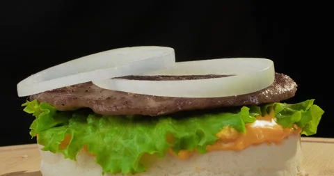 Delicious hamburger or cheeseburger preparation. Onion rings fall on bun with Stock Footage 293323125
