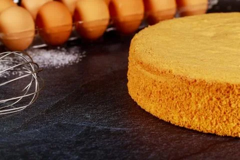 Delicious sponge cake on dark table with raw eggs. Bakery concept. Stock Photos