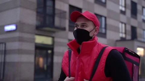 Delivery guy wearing red uniform and protective mask while walking along modern Stock Footage 144192842