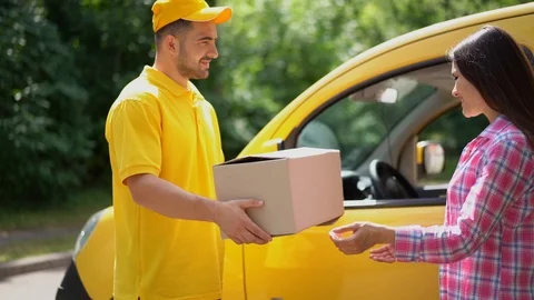 Delivery guy in yellow uniform gives parcel to female customer near car Stock Footage 99697293