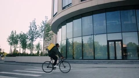Delivery man on a bicycle with a yellow backpack walks along an office building Stock Footage 217363093