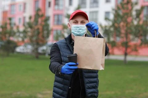 Delivery man in red cap, face medical mask, gloves hold take away paper bag a Stock Photos