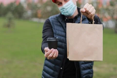 Delivery man in red cap, face medical mask hold take away paper bag and drink in Stock Photos