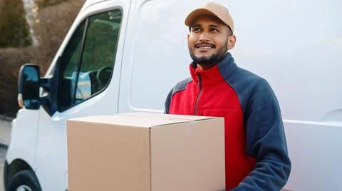 Delivery man smiling while holding parcel in front of transportation van Stock Photos