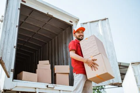 Delivery man smiling while lifting parcel box against container background Stock Photos