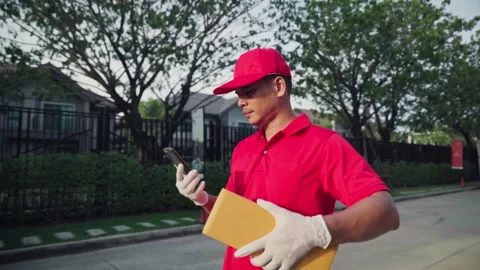 A delivery man wearing a red shirt walks around holding a parcel box and using h Stock Footage 245362353