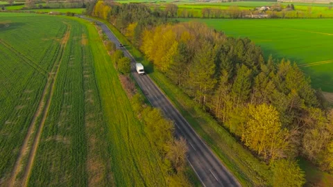 Delivery van moving along rural road between green fields and forest in spring Stock Footage 333255607