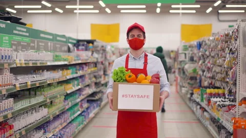 Deliveryman in face mask and uniform standing in supermarket with vegetables in Stock Footage 147994862