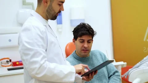 Dentist showing digital tablet to patient. He is sitting in the dentist's cha Stock Footage 295626527