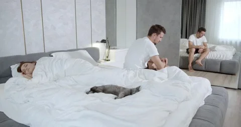 Depressed impotent man is sitting on the edge of the bed, woman is sleeping, 4k Stock Footage 135376341