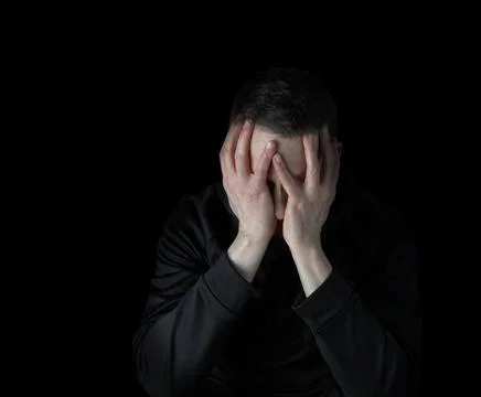 Depressed man with face down in both hands while surrounded by darkness Stock Photos