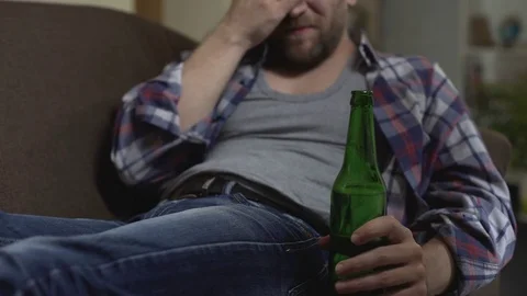 Depressed man sitting on couch drinking beer, scratching belly, money problems Stock Footage 84875960