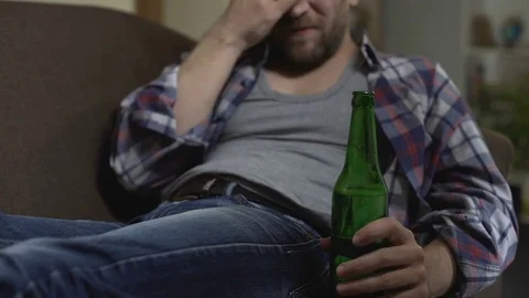 Depressed man sitting on couch drinking beer, scratching belly, money problems Stock Footage 84890990