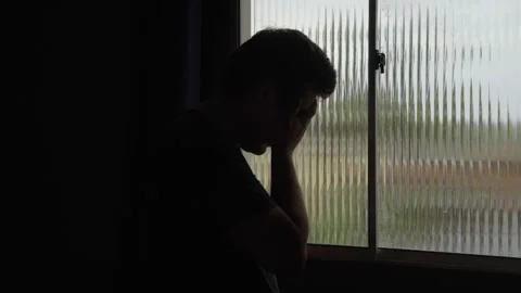 Depressed man standing in front of a window, covering his face with his hands  Stockbeeldmateriaal 329655657