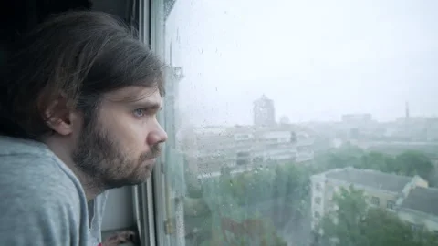 Depressed sad man stands at the window on rainy day at view of city skyline Stock Footage 277646598