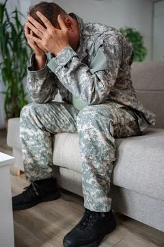 Depressed soldier sitting on sofa holding head in hands Stock Photos