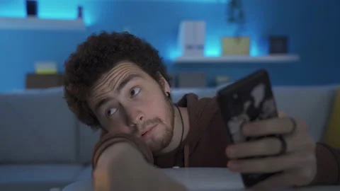 Depressed young man looking at his phone at home bored and unhappy. 库存影片 233016038