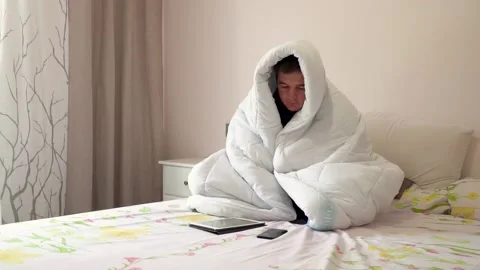 A depression man sits on a bed wrapped in a blanket with a tablet and a phone Stock Footage 132040685