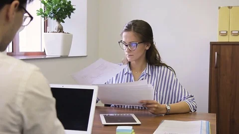 At desk woman asks question on documents to young man who sits opposite. Stock Footage 82235402