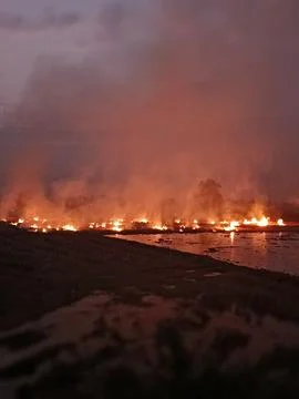 Destructive Field Fire and Glowing Flames Reflected on Water Surface at Dusk Stock Photos