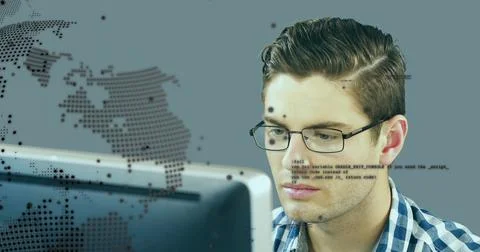 Developer wearing glasses reviewing code on monitor at desk, with world map o 库存照片