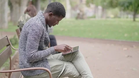 Device, accessories - black man sitting on a park bench types on tablet Stock Footage 240392556