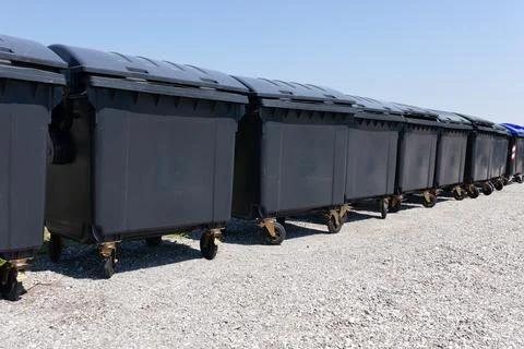 Diagonal row of black garbage containers on gravel outside under the blue sky Foto stock