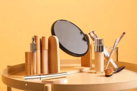 Different cosmetics, makeup brushes and mirror on table near yellow wall in l Stock Photos