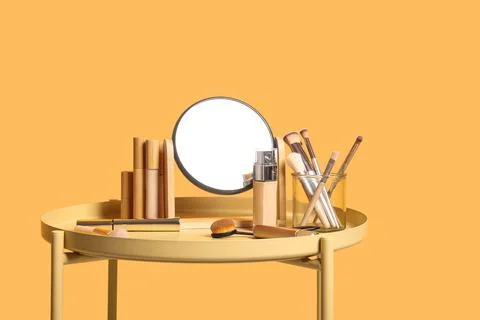 Different cosmetics, makeup brushes and mirror on table near yellow wall in l Stock Photos