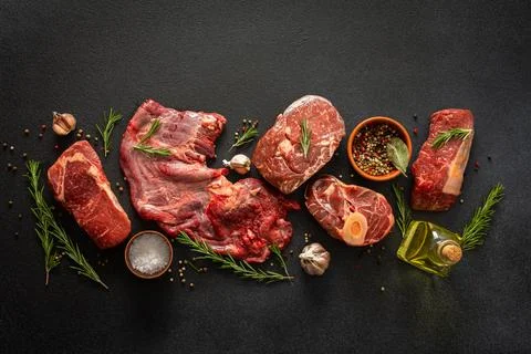 Different cuts of beef steak are arranged on a black surface. Fresh herbs, .. Stock Photos