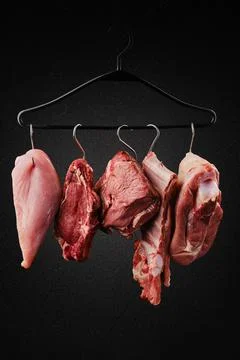 Different cuts of raw meat on hook on hanger over black background Foto stock
