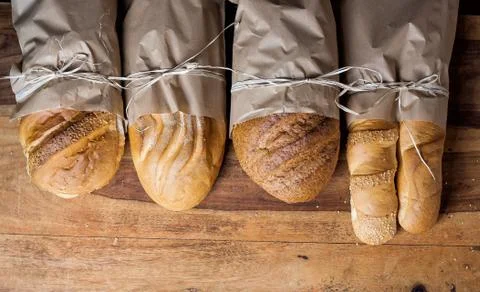 Different fresh baked bread in paper bags on wooden table. Stock Photos