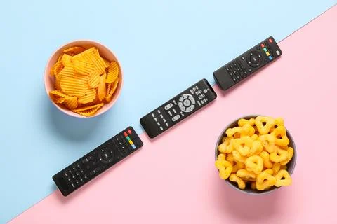 Different modern TV remote controls with bowls of crispy snacks on color back Stock Photos