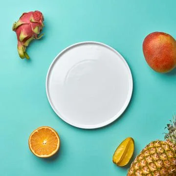 Different tropical fruits and empty white plate on a blue background with c.. Stock Photos