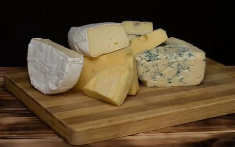 Different types of cheese on a rustic wooden cutting board on a dark backgrou Stock Photos