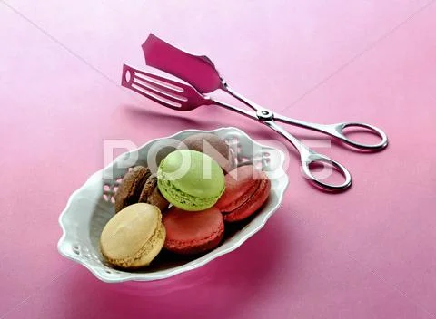 Different types of macaroons in a porcelain dish with a pair of tongs next to it Stock Photos