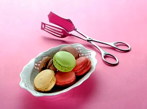 Different types of macaroons in a porcelain dish with a pair of tongs next to it Fotos Stock