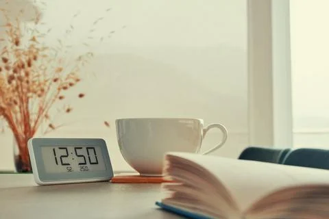 Digital clock, white coffee cup and an opened book resting on the table Stock Photos