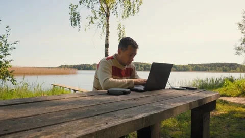 Digital nomad working remotely at a lakefront picnic table Stock Footage 322650791
