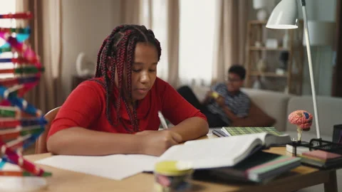 Diligent girl doing homework while her brother plays video games on background Stock Footage 334947514