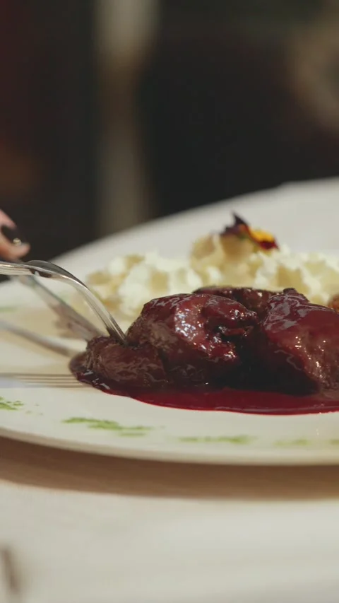 A diner uses a fork and knife to cut cooked meat coated in glossy red sauce on Stock Footage 348687951