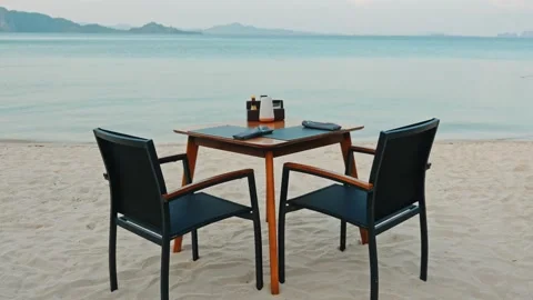 Dining table and chairs on sandy beach, ocean background, resort 動画素材 334411590