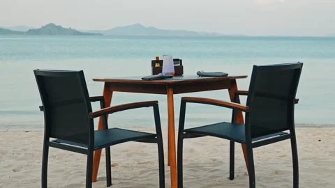 Dining table and chairs set on sandy beach facing ocean, resort travel Stock Footage 340555440