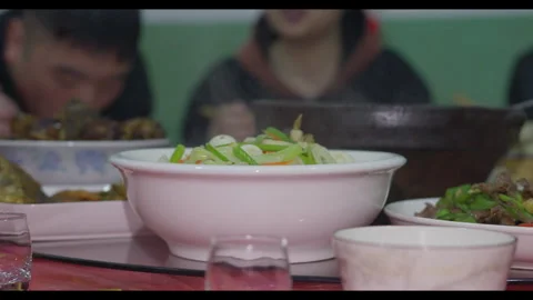 Dining table with multiple dishes, people eating in background, no faces Stock Footage 329695702