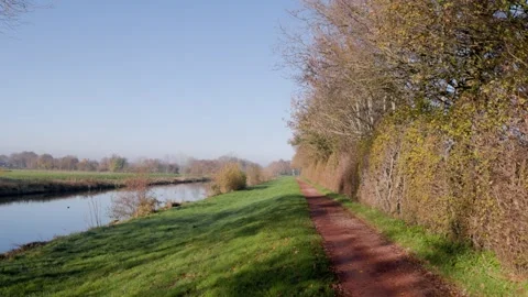 Dirt path beside calm river bordered by leafless trees and grassy bank clear Vídeos de archivo 323359649