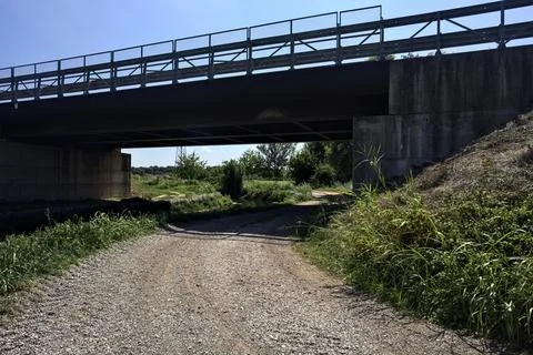 Dirt path next to a brook bordered by trees and reeds with a viaduct passin.. Stock Photos